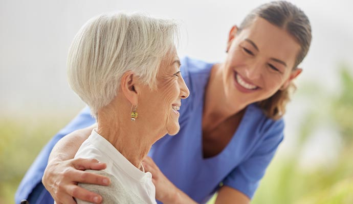 A smiling female caregiver in blue scrubs gently placing her hands on the shoulders of an elderly woman