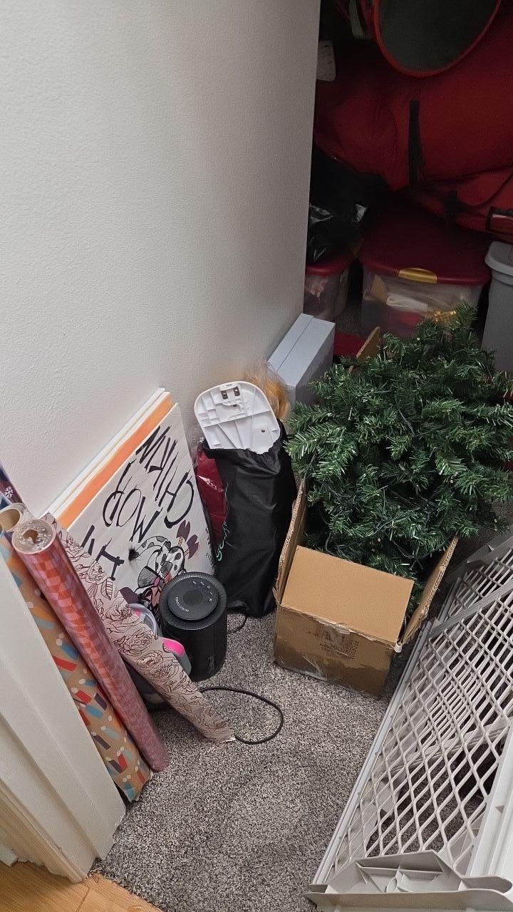 A cluttered closet space with various items including a Christmas tree box and a small black fan on a carpeted floor, suggesting a drying process during water damage restoration.